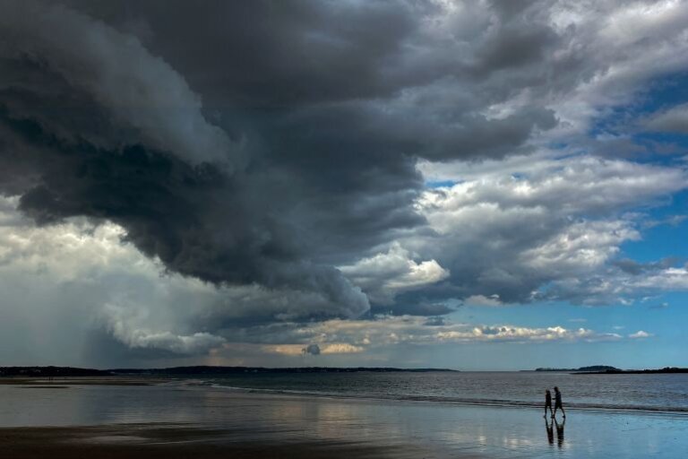 People walk under dark clouds along a beach in the United States in 2023 [File: Kevin Lamarque/Reuters]